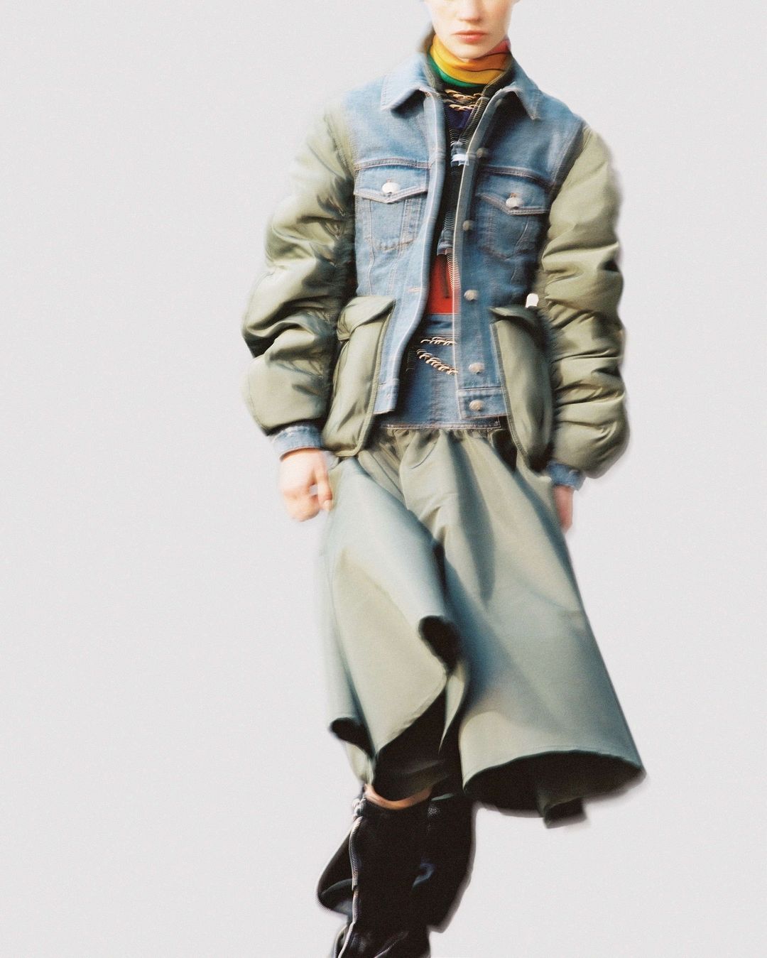 Editorial fashion shot of an adult woman wearing a patchwork denim and puffer jacket with a long olive skirt against a light background.