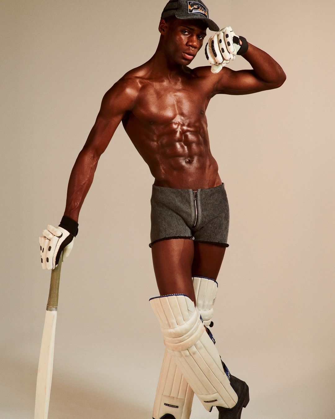 Athletic Black man in cricket gear poses in a studio setting with bat and gloves, showing a fit physique and confident stance.