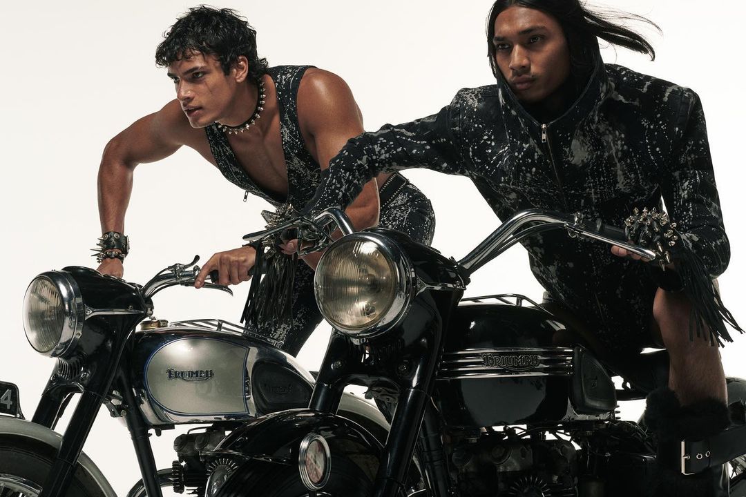 Two male models pose with Triumph motorcycles against a bright backdrop. The motorcycles are black and silver, with visible logos.