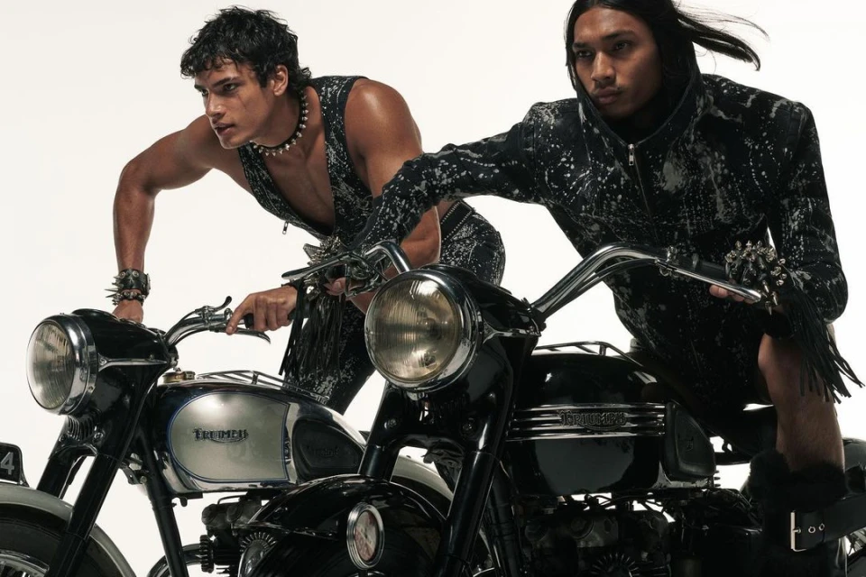 Two male models pose with Triumph motorcycles against a bright backdrop. The motorcycles are black and silver, with visible logos.
