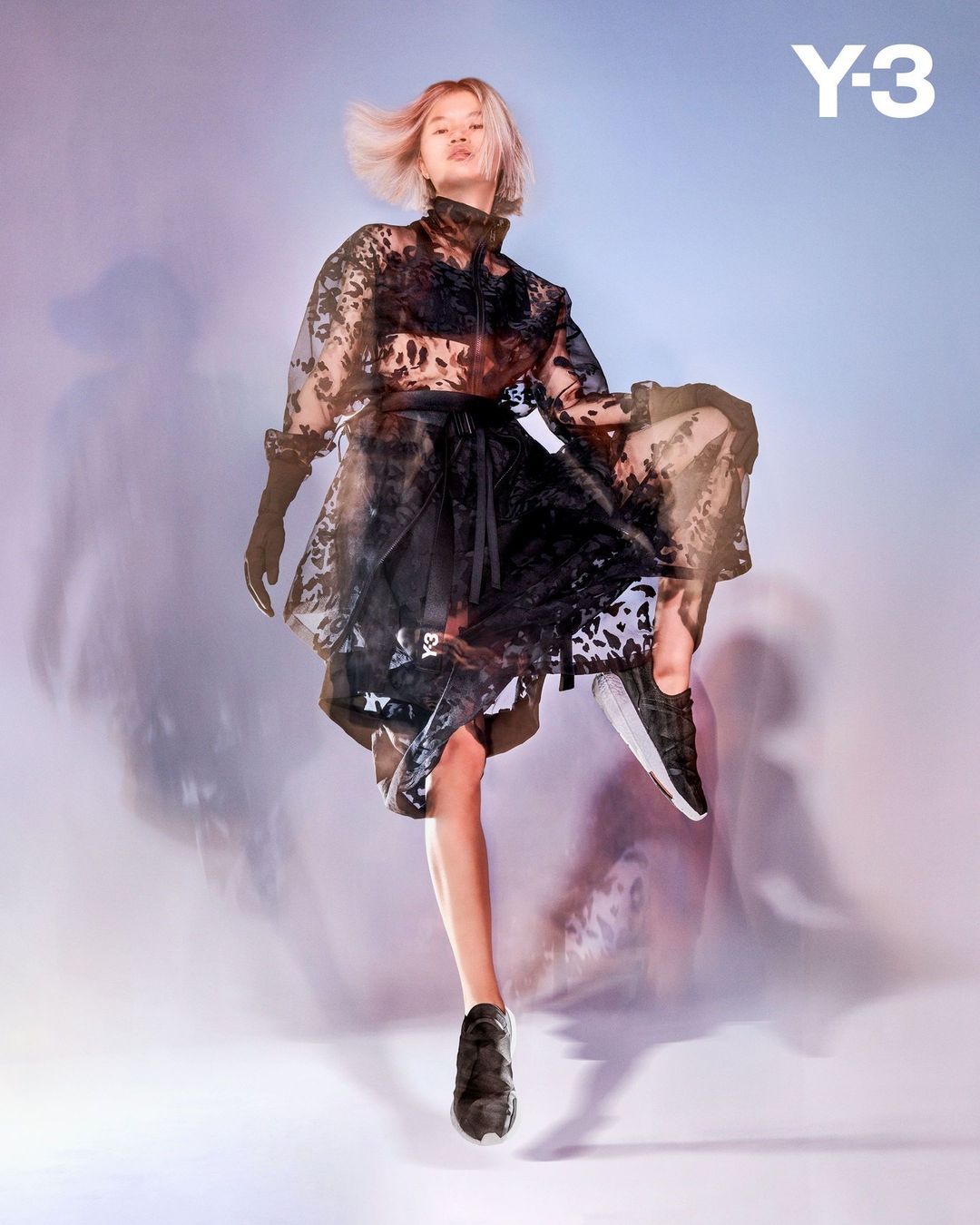 Fashion shot of a blonde woman in a sheer black outfit and athletic shoes mid-air against a gradient background with the Y-3 logo.