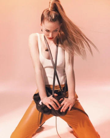 Woman in white tank top and orange trousers holding a black handbag against a warm gradient background by Heji Shin.