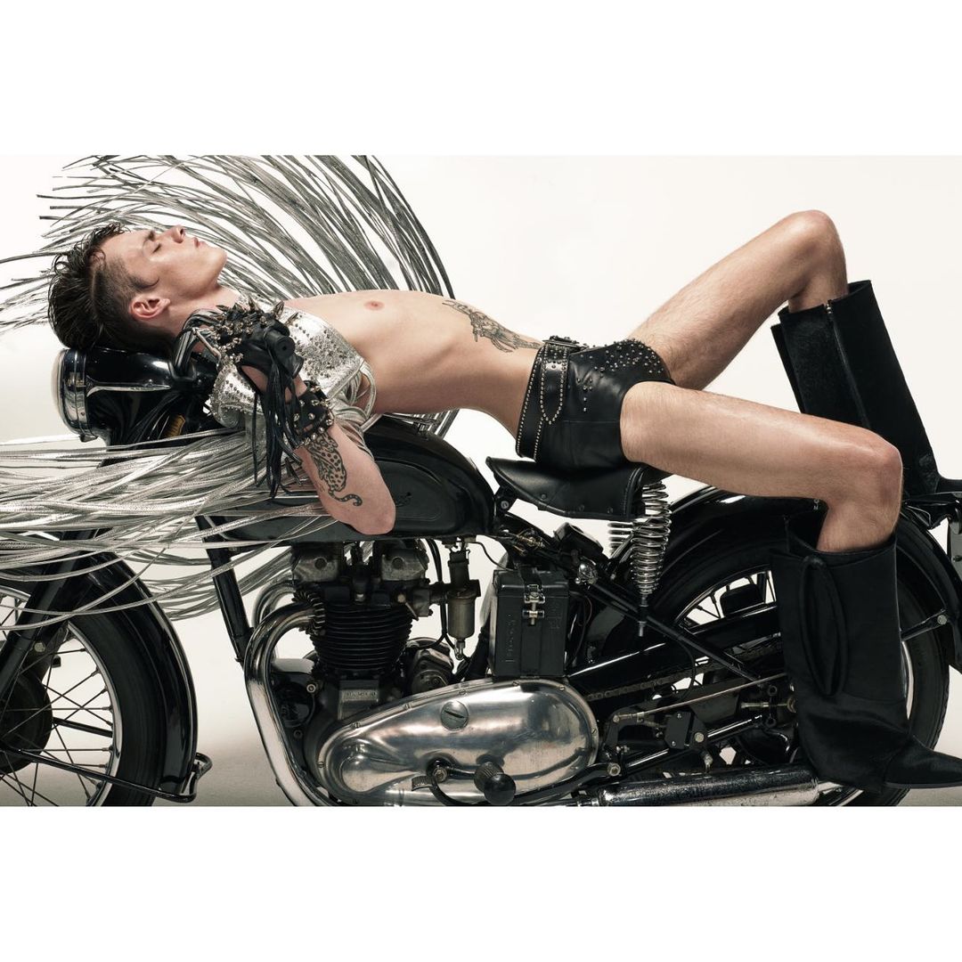 Shirtless adult male model reclines on a black motorcycle with chrome accents in a studio. He wears leather shorts and black boots.