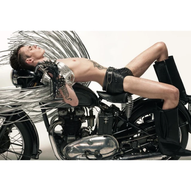Shirtless adult male model reclines on a black motorcycle with chrome accents in a studio. He wears leather shorts and black boots.