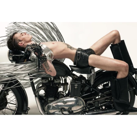 Shirtless adult male model reclines on a black motorcycle with chrome accents in a studio. He wears leather shorts and black boots.