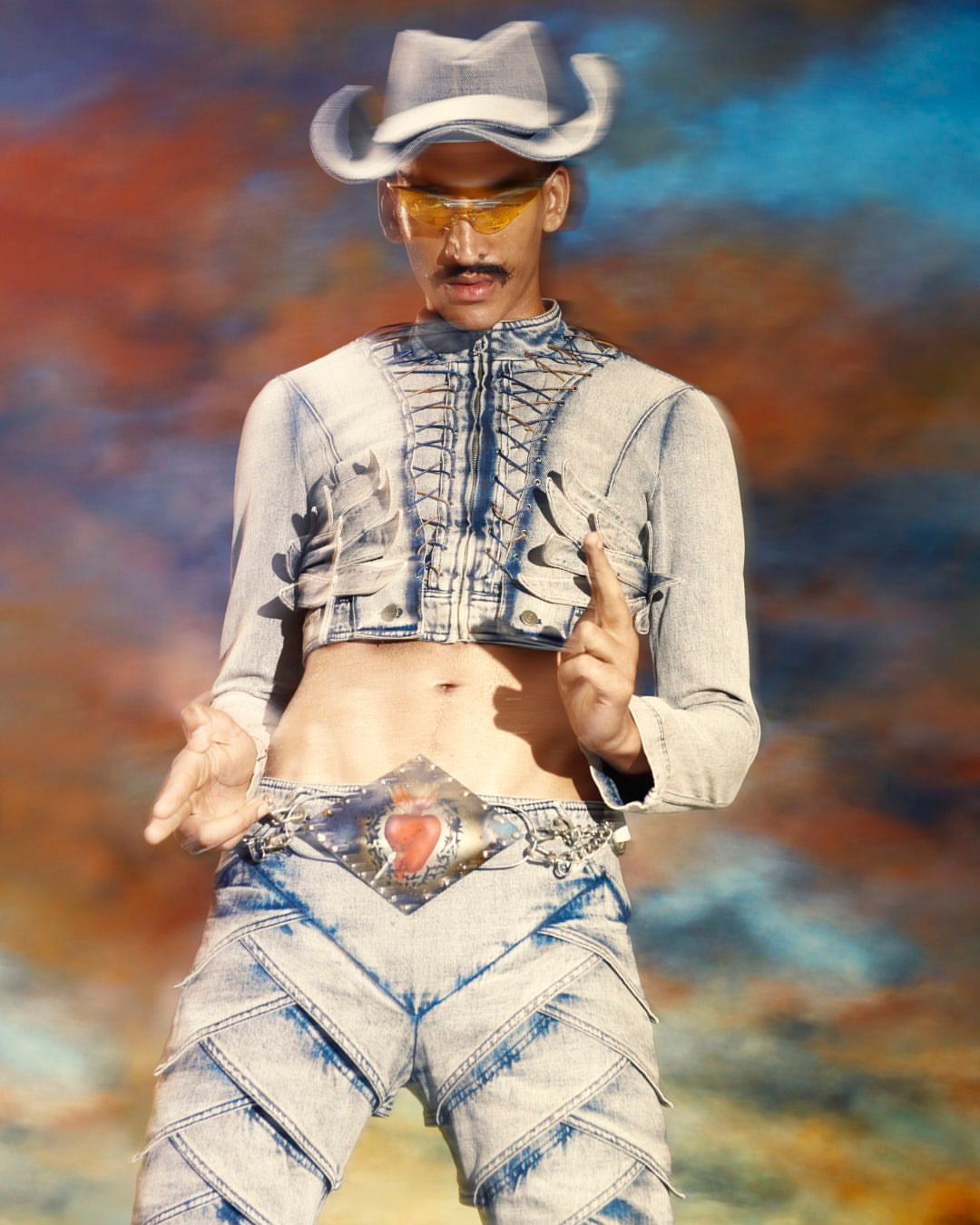 Man with mustache poses in light-blue cropped denim jacket, jeans, matching cowboy hat and orange sunglasses against a colorful background.