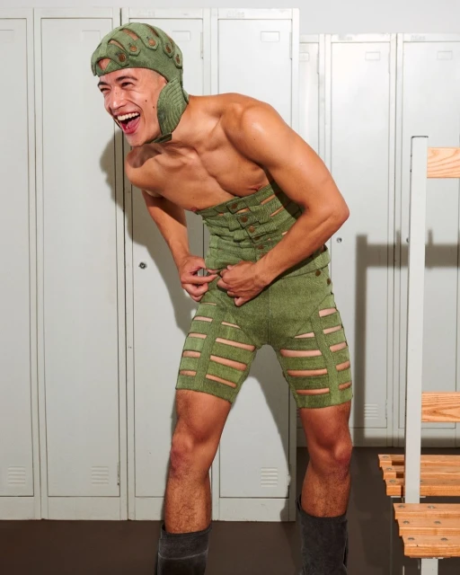 Laughing man in a green textured outfit with a hat poses in front of metal lockers, showcasing a unique and eye-catching design.