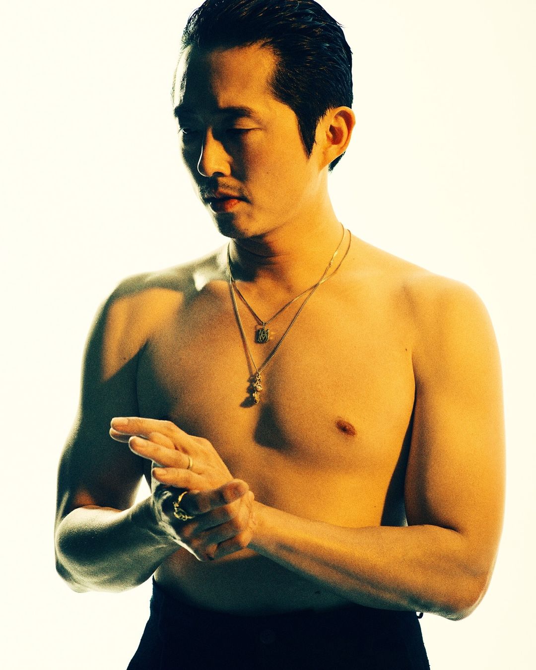Shirtless Asian man with gold necklaces and rings stands indoors against a white background, posing while touching his hands together.