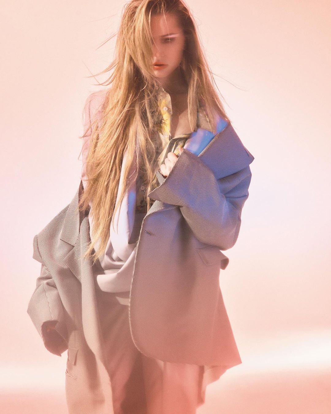 Fashion shot of a young woman with long blonde hair wearing a grey jacket, styled in a studio with soft, diffused pink and blue light.