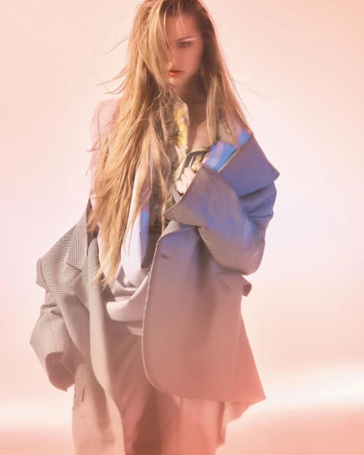 Fashion shot of a young woman with long blonde hair wearing a grey jacket, styled in a studio with soft, diffused pink and blue light.