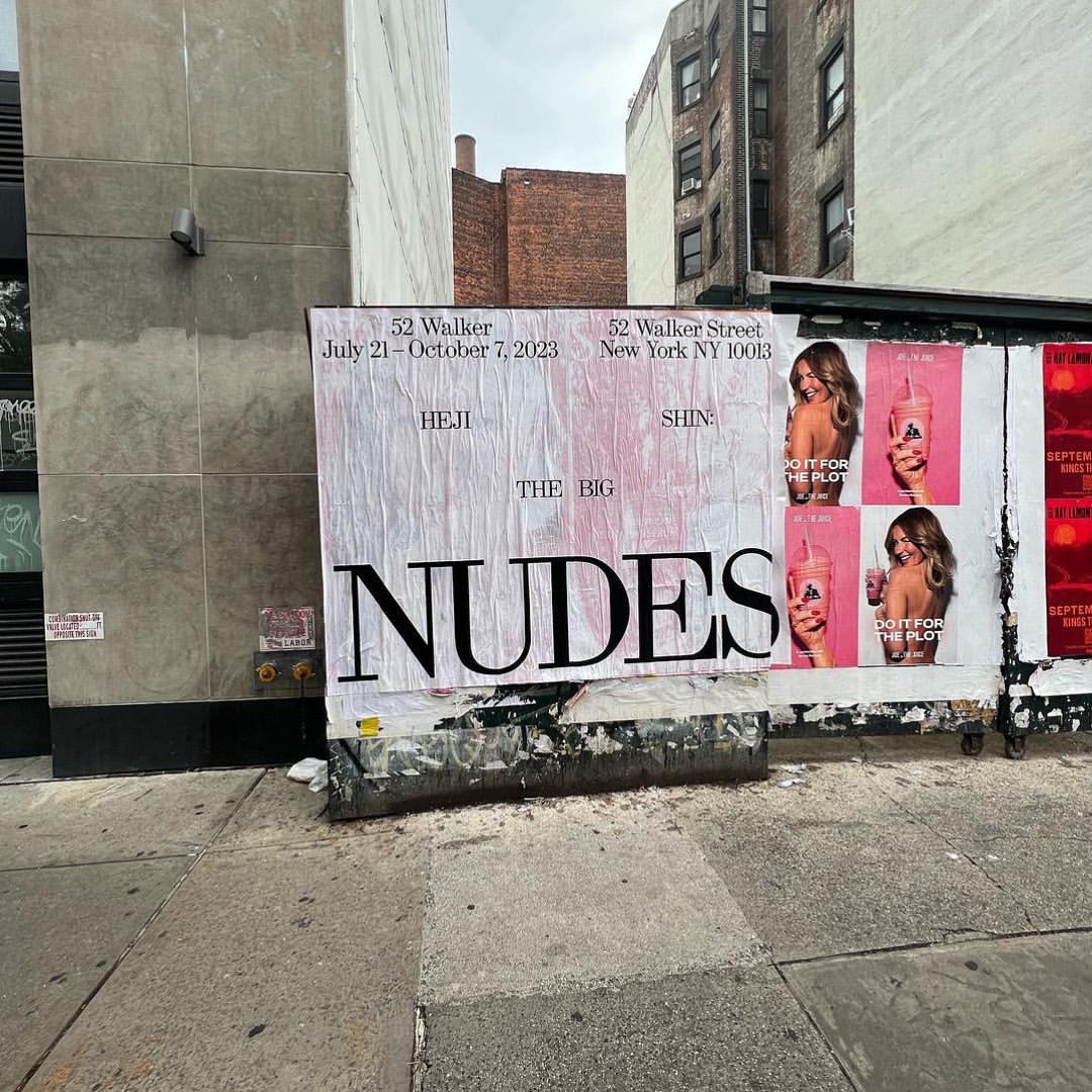 Outdoor billboard on a city street shows posters for a gallery show called The Big Nudes with pink ads featuring a woman holding a drink.