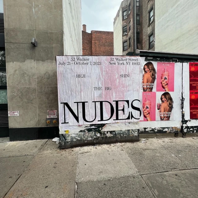 Outdoor billboard on a city street shows posters for a gallery show called The Big Nudes with pink ads featuring a woman holding a drink.