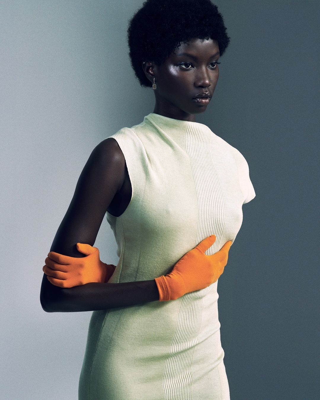 Elegant portrait of a Black female model in a form-fitting light-green dress and bright orange gloves, posing in front of a grey background.