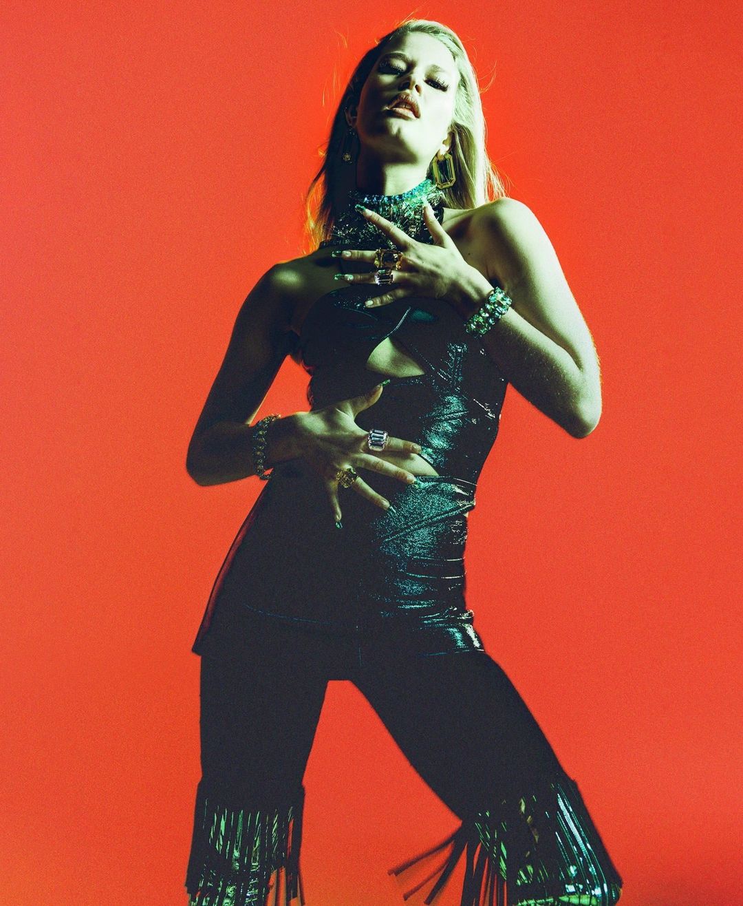 Fashionable young woman poses in a shiny black outfit with fringe, jewelry against a solid orange backdrop in a studio shot.