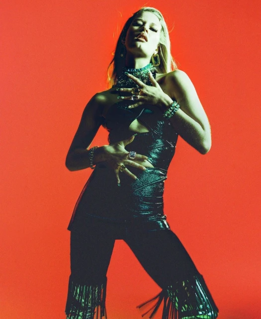 Fashionable young woman poses in a shiny black outfit with fringe, jewelry against a solid orange backdrop in a studio shot.