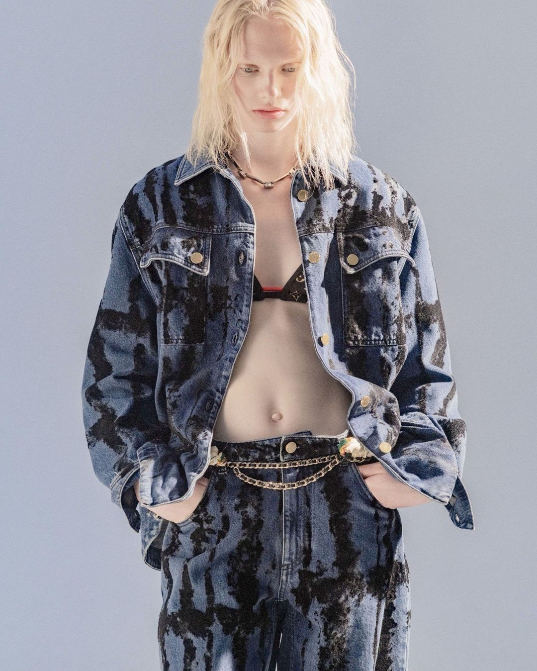 A person with long blonde hair poses in matching denim jacket and jeans with black tie-dye pattern and gold chain belt against light-blue backdrop.