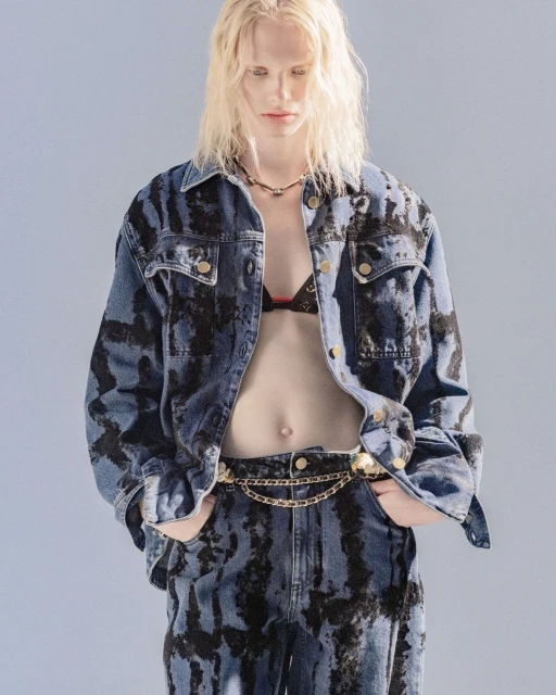 A person with long blonde hair poses in matching denim jacket and jeans with black tie-dye pattern and gold chain belt against light-blue backdrop.