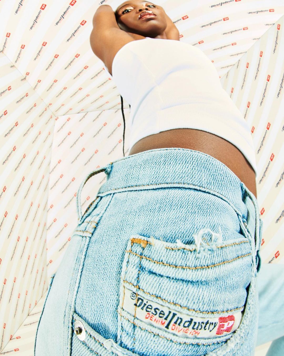 Woman in jeans and white top, low-angle shot, indoors with patterned white backdrop. Denim pocket with logo in view. Fashion and beauty.