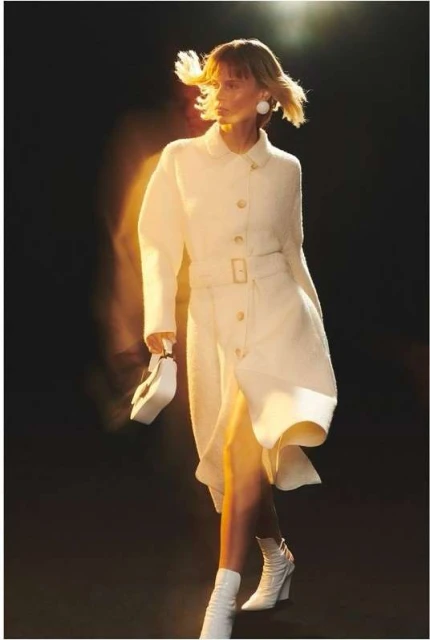 Fashion model in a belted, long white coat dress with matching accessories walks in a dark studio with diffused lighting.