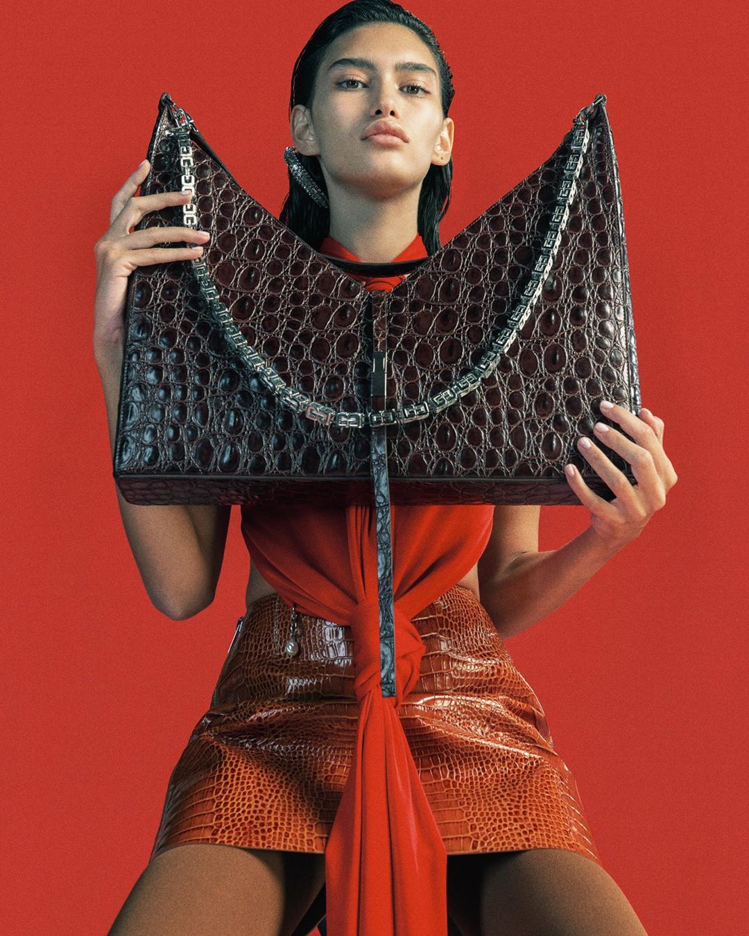 Woman in orange skirt poses with oversized dark brown crocodile-skin purse against red backdrop in fashion photography studio shot.