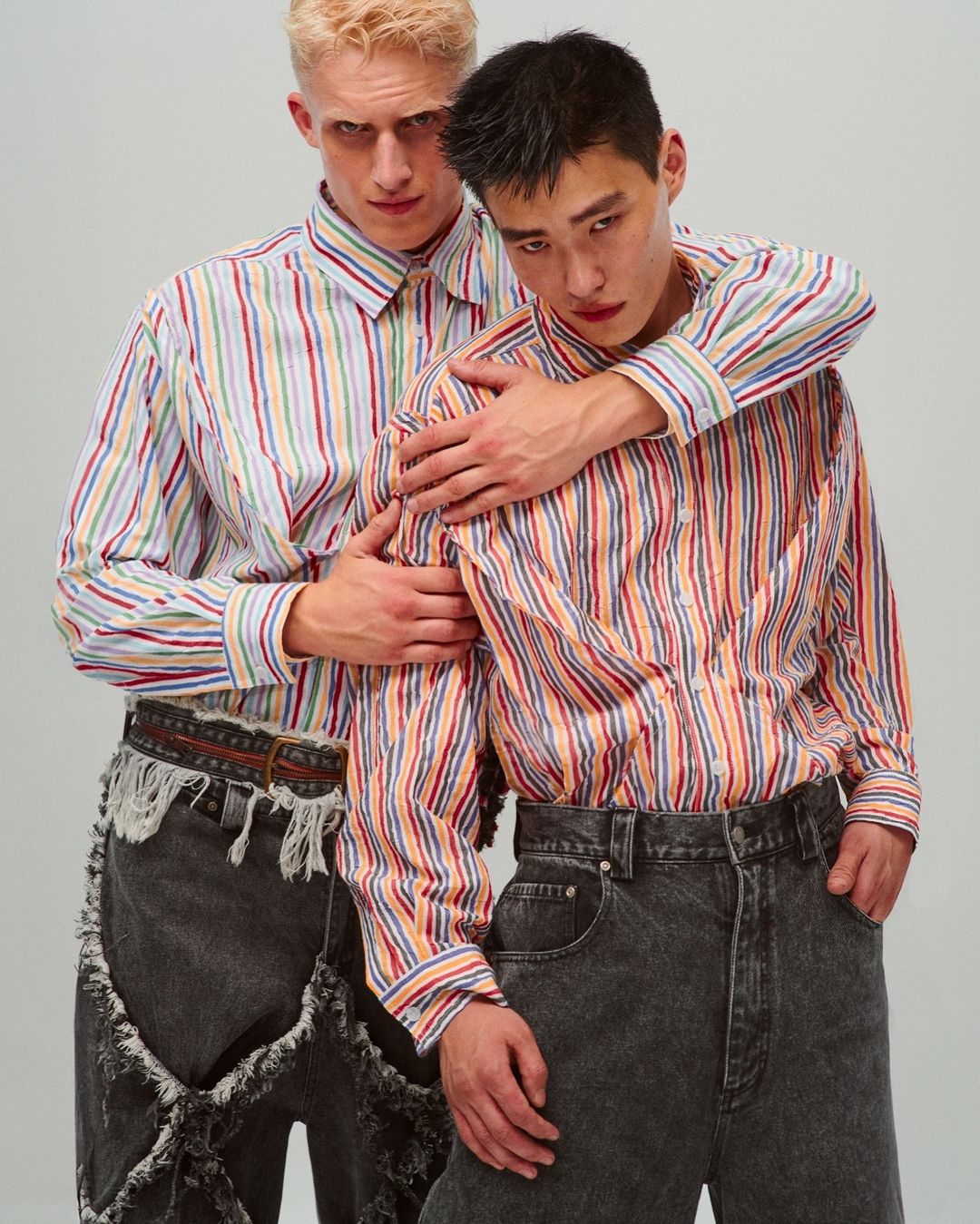 Two young men in striped shirts and acid wash jeans pose in a studio setting, one with blond hair and the other with dark hair.