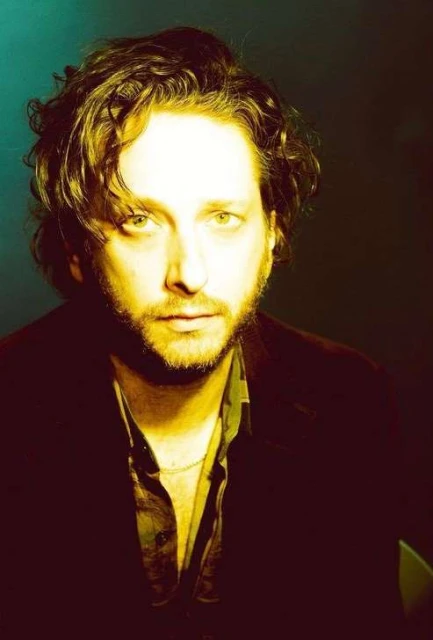 Close-up portrait of a man with curly brown hair, a beard, and a dark jacket against a dark green background, with golden-toned lighting.