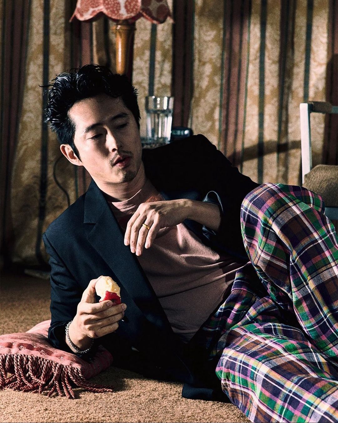 Steven Yeun in jacket and plaid pants lying on floor with lamp and drink in background holding an apple in eye-level shot.