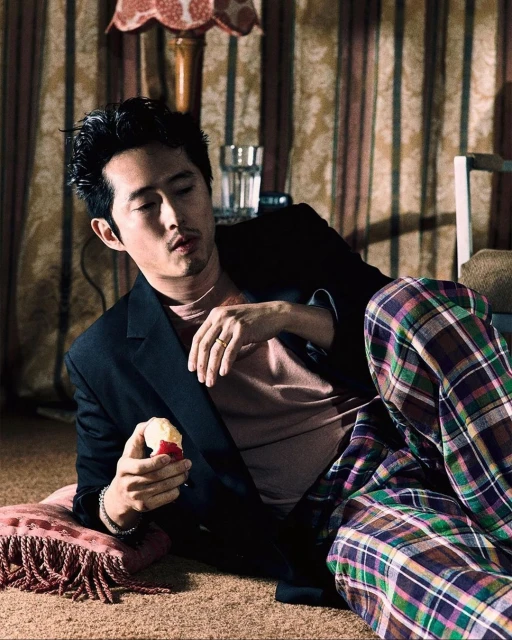 Steven Yeun in jacket and plaid pants lying on floor with lamp and drink in background holding an apple in eye-level shot.