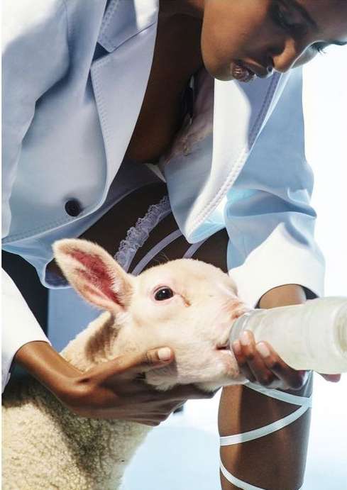 Eye-level shot of woman in a white jacket feeding a lamb from a clear plastic bottle, with a light blue background and bright studio lighting.