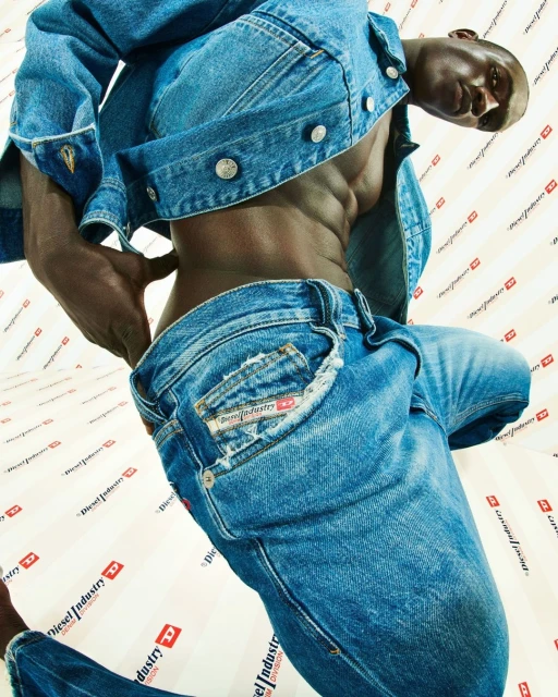 A muscular black man wears a denim jacket and jeans with a Diesel logo background, showcasing a fashion-forward look.