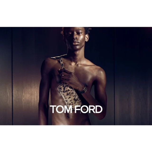 A shirtless dark-skinned male fashion model with a leopard-print cross-body bag poses in front of a dark background in a studio shot.