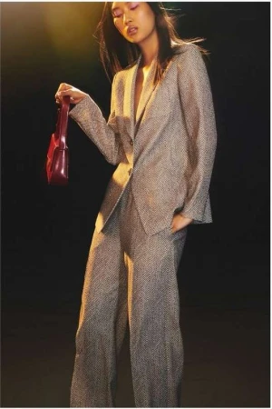 Asian woman in a patterned grey suit holds a red purse against a black background in a fashion-style full shot.