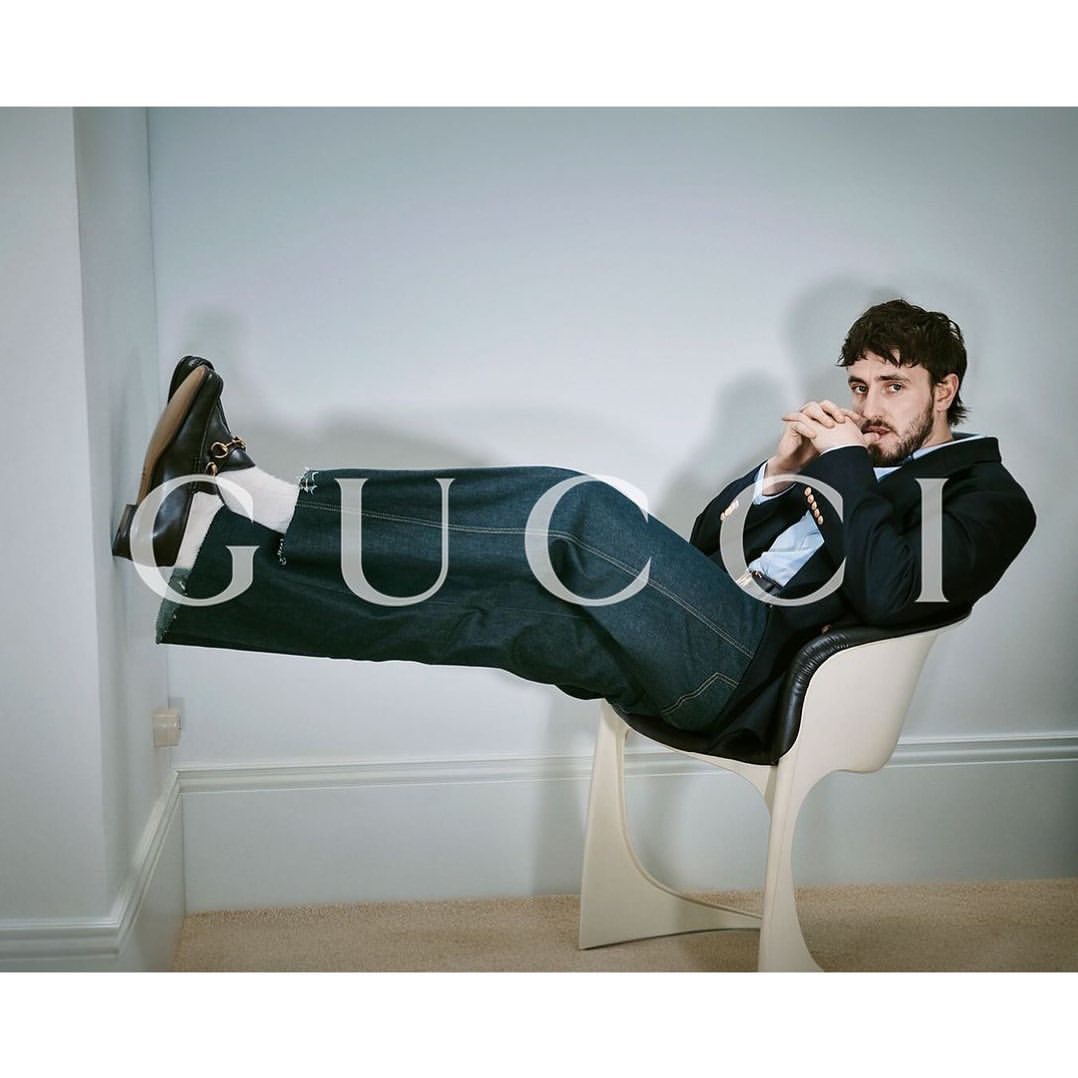 Man in dark blazer and jeans seated in a white chair with legs propped horizontally against a wall, shot by Heji Shin.