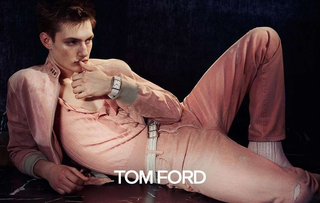 A fair-skinned adult male fashion model reclines in pink denim and jacket, accessorized with a white watch, in a studio shot.