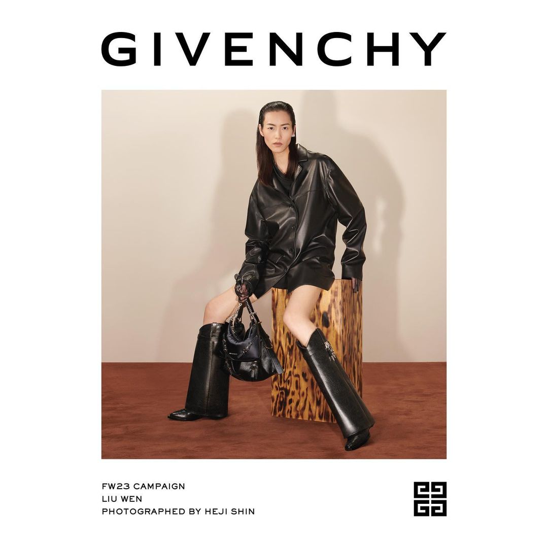 An adult Asian woman models a black leather jacket dress, high boots, and handbag for Givenchy, seated indoors against a neutral backdrop.