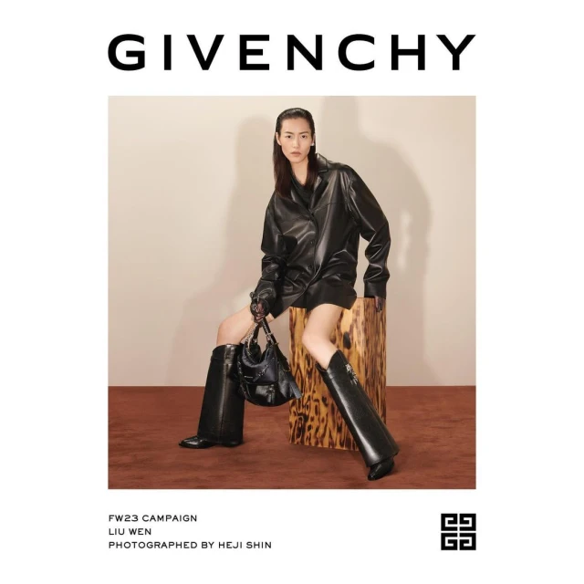 An adult Asian woman models a black leather jacket dress, high boots, and handbag for Givenchy, seated indoors against a neutral backdrop.
