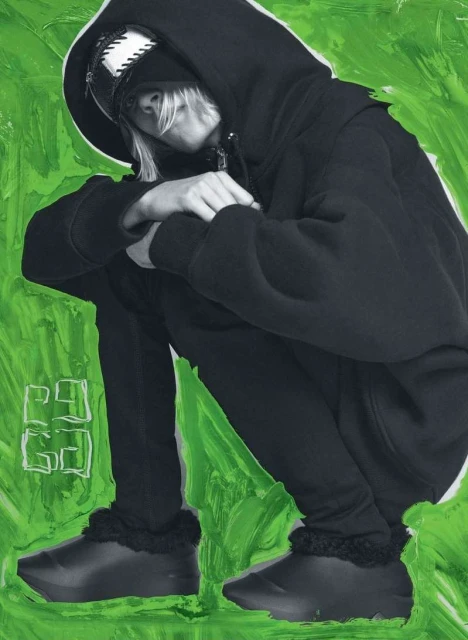Young adult squats in black hooded sweatshirt and pants against a green paint-stroke background in a stylized portrait.
