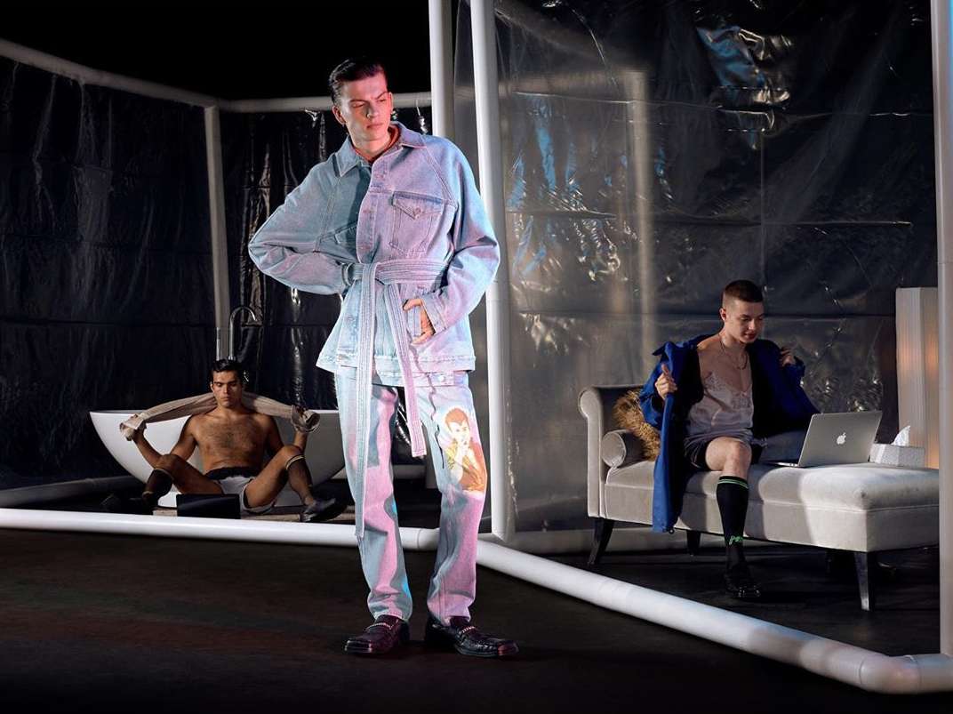 Fashion shoot featuring a male model in a denim outfit, another in a bathtub, and a third on a chaise lounge with a laptop, in studio.