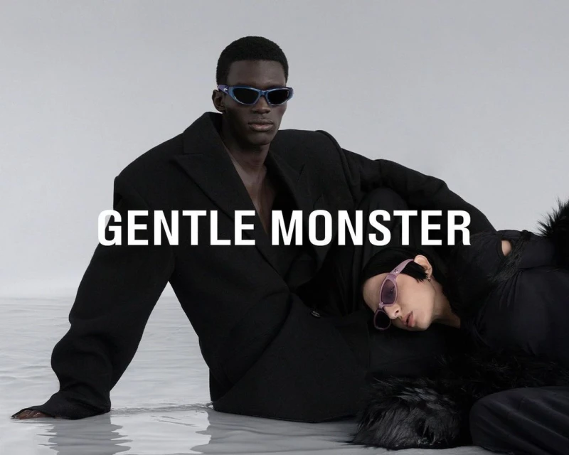 Two fashion models, one lying down, and the other sitting in a black blazer, wear trendy sunglasses in a studio shot.