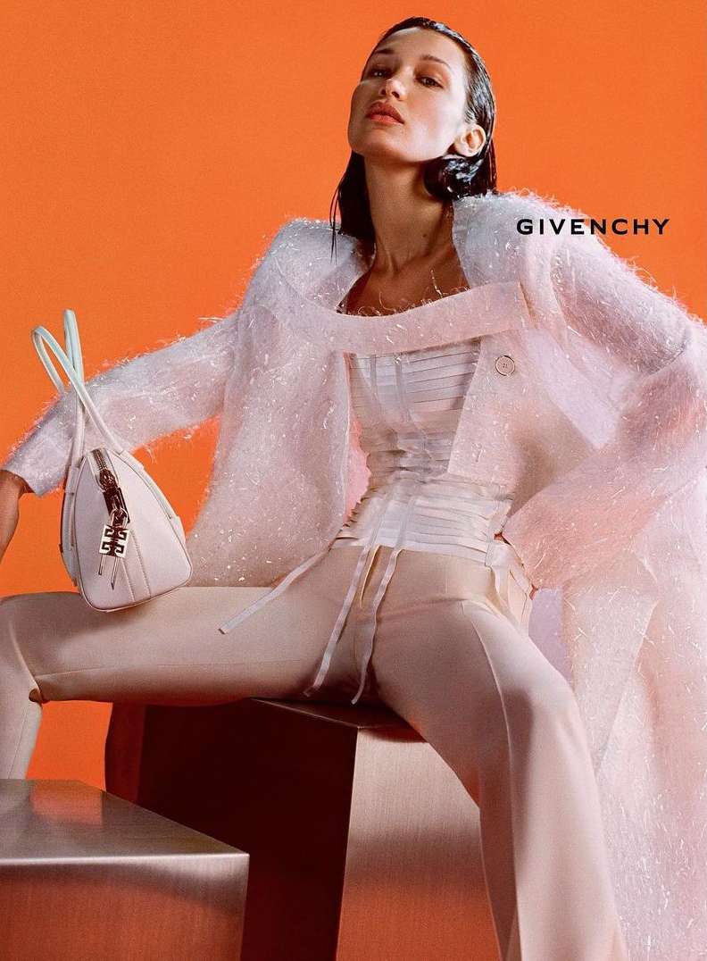 Fashion editorial shot of an adult female model in a white jacket and cream pants, holding a handbag, posing against an orange backdrop.