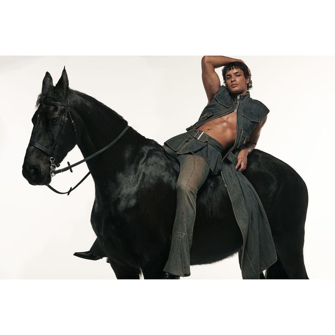 A muscular adult male wearing a jean vest and trousers poses bare-chested on a shiny black horse against a seamless white backdrop.