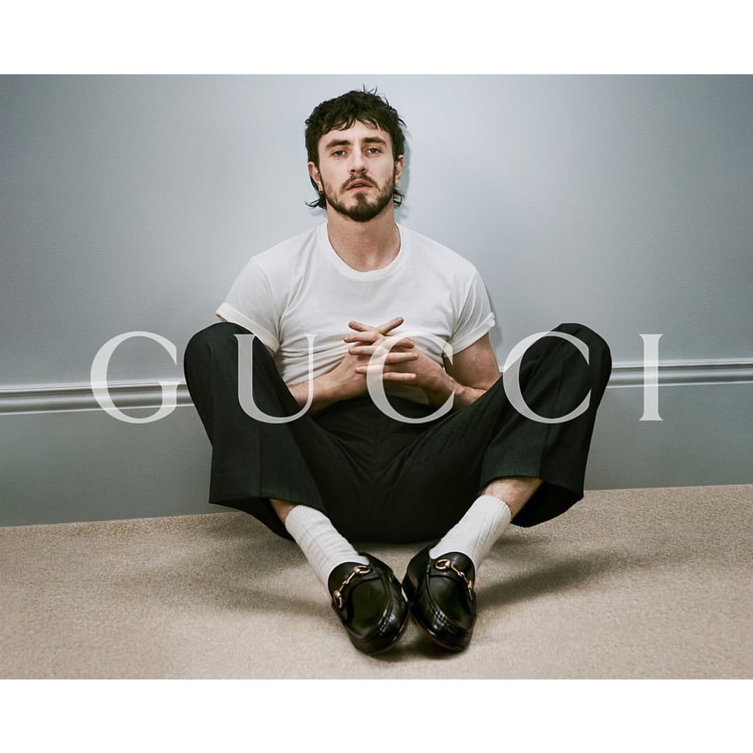 Man in white t-shirt and black pants sits on beige carpet with black Gucci loafers, positioned in front of a gray wall with white trim.