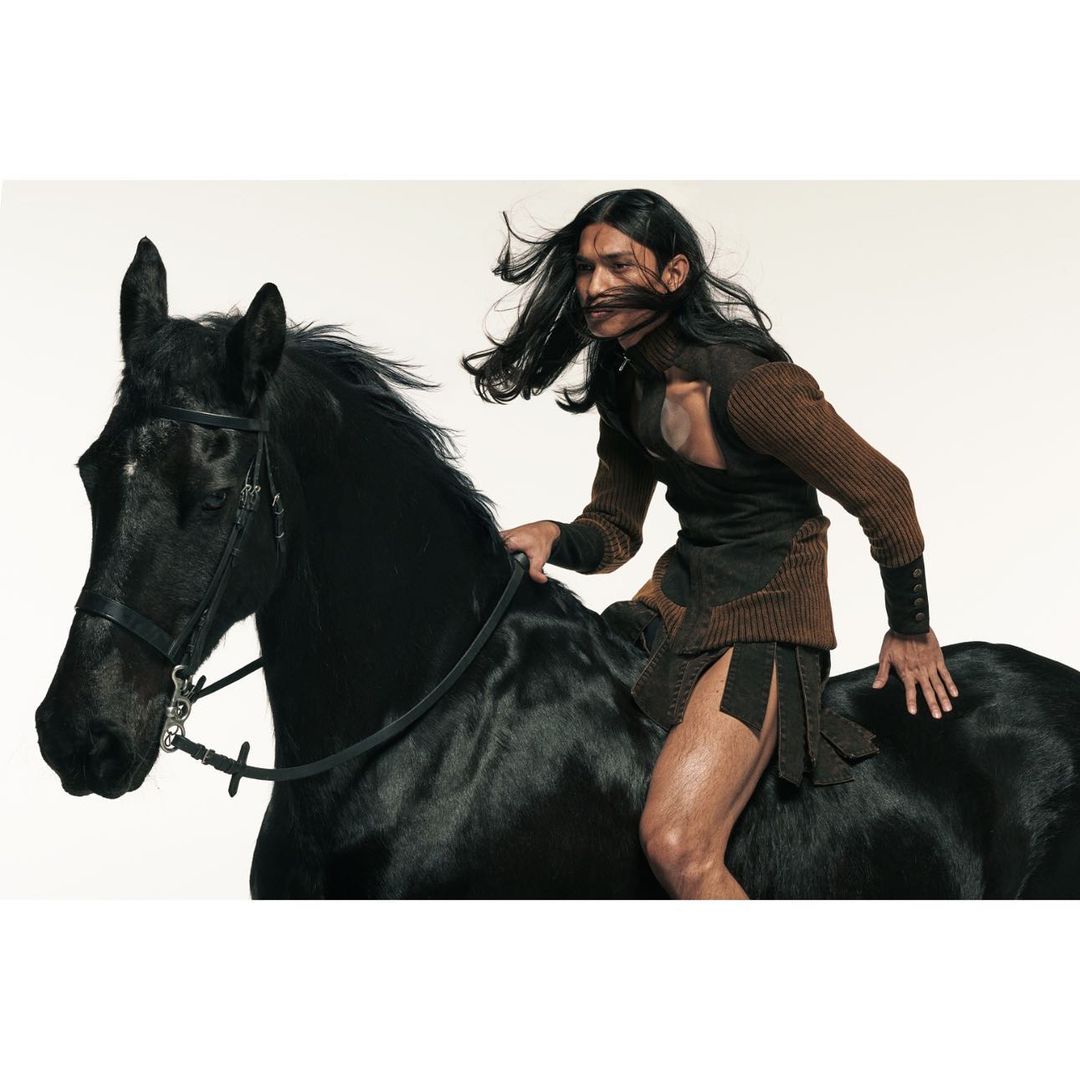 A man with long dark hair riding a black horse in a photo studio. The man is wearing an unusual outfit. The horse has a harness.