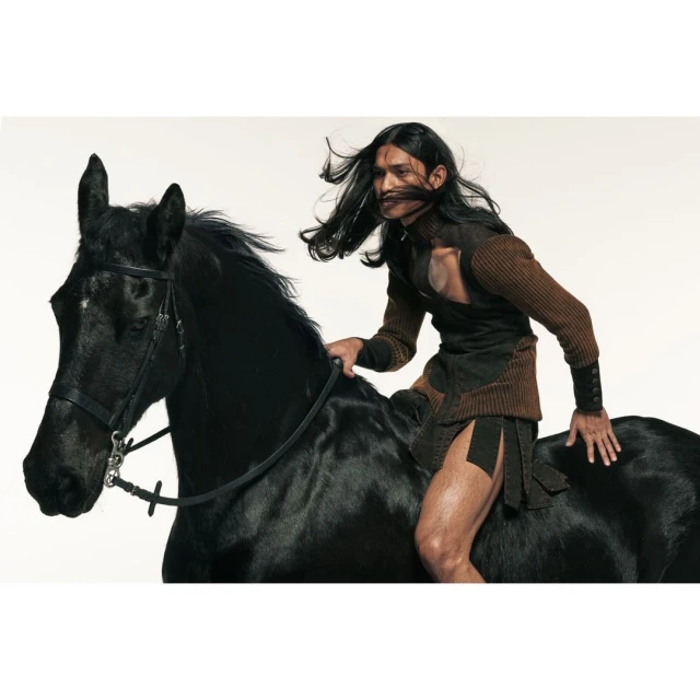 A man with long dark hair riding a black horse in a photo studio. The man is wearing an unusual outfit. The horse has a harness.