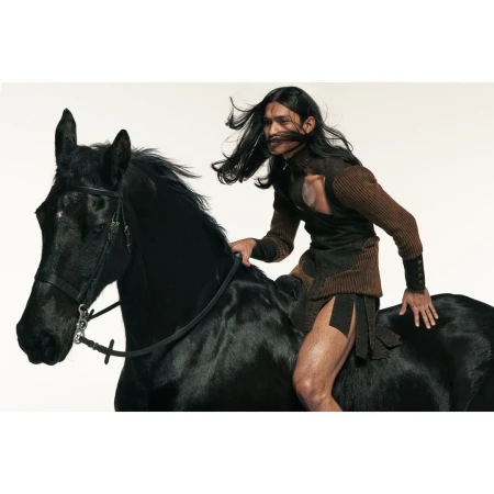 A man with long dark hair riding a black horse in a photo studio. The man is wearing an unusual outfit. The horse has a harness.