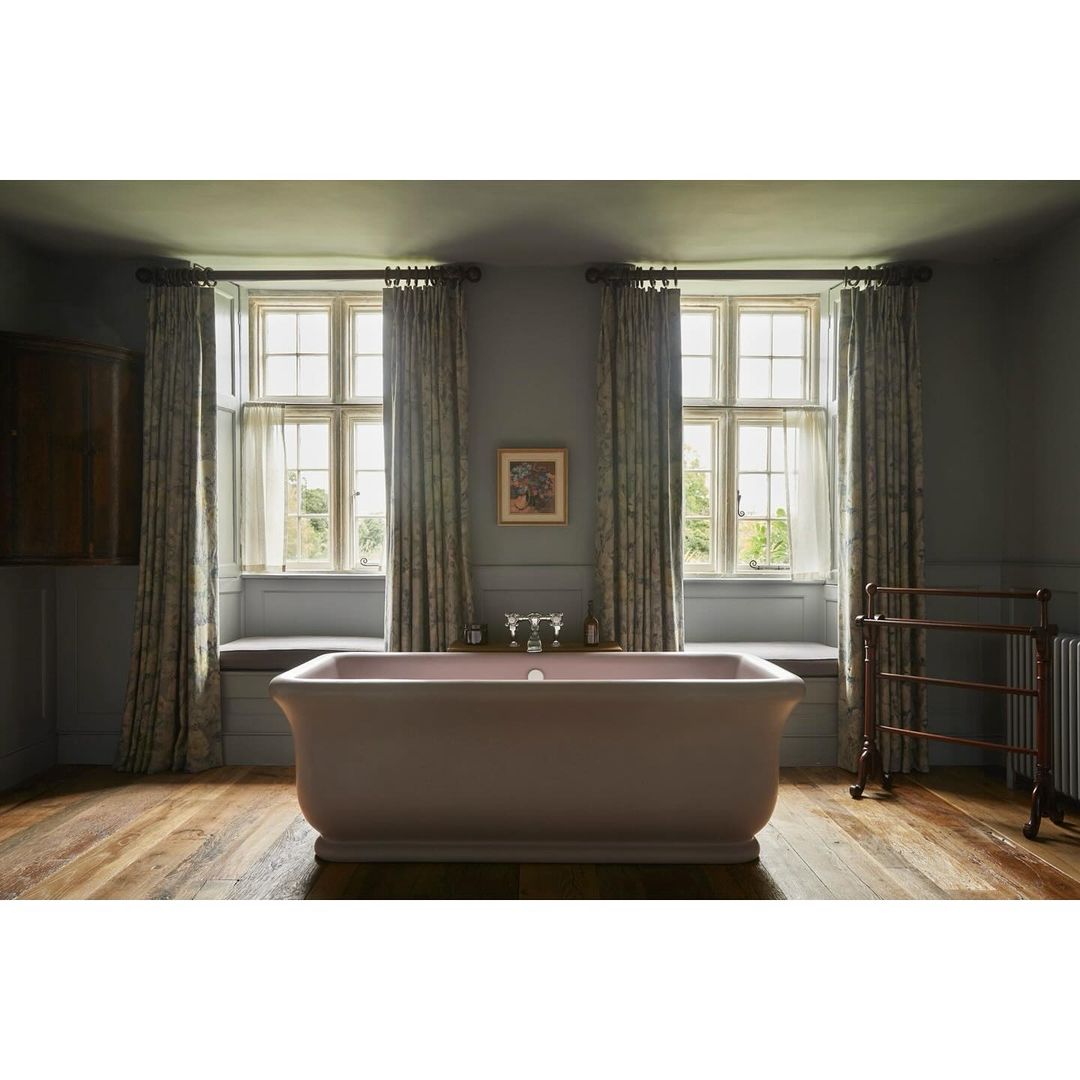 Bathroom interior featuring a standalone light pink bathtub on a wood floor, flanked by windows with patterned curtains and a radiator.