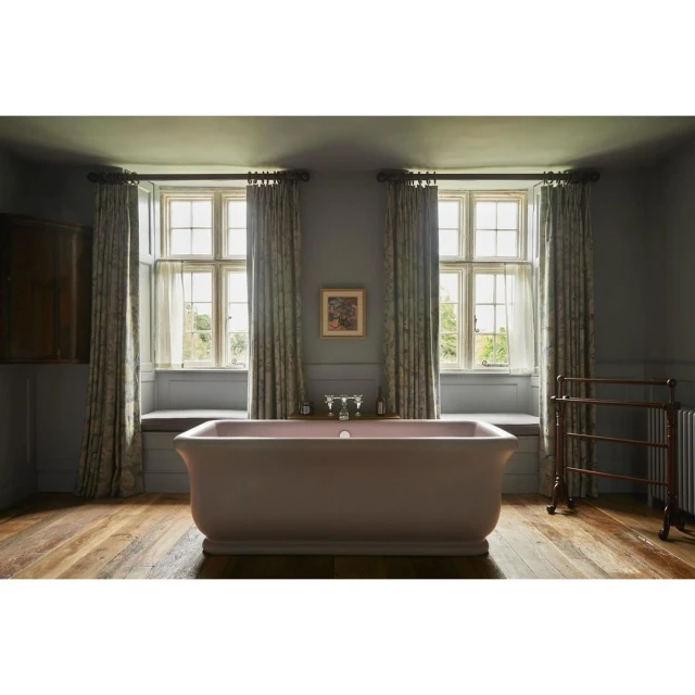 Bathroom interior featuring a standalone light pink bathtub on a wood floor, flanked by windows with patterned curtains and a radiator.