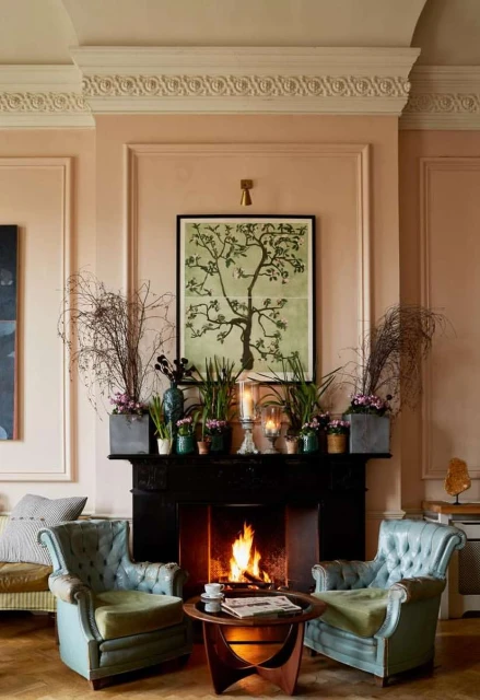 Living room interior featuring a black fireplace with a burning fire, flanked by two blue armchairs and decorated with plants and artwork.
