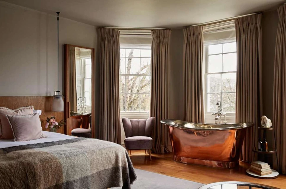 Bedroom and bathroom interior with a copper bathtub near a window with curtains, a bed, an armchair, and neutral color tones.
