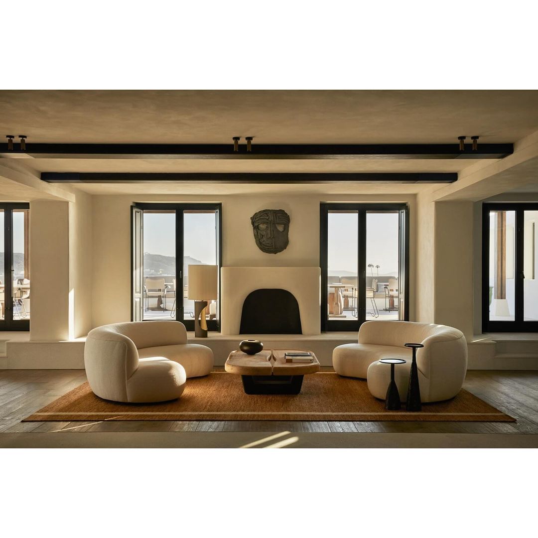 Living room interior featuring two curved sofas, a central coffee table, fireplace, and large windows with a view of a coastal landscape.